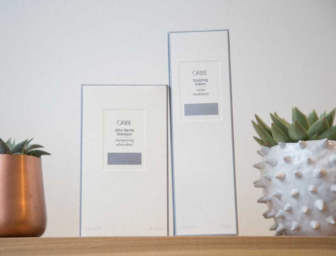 Oribe shampoo and cream boxes on a shelf with decorative plants.